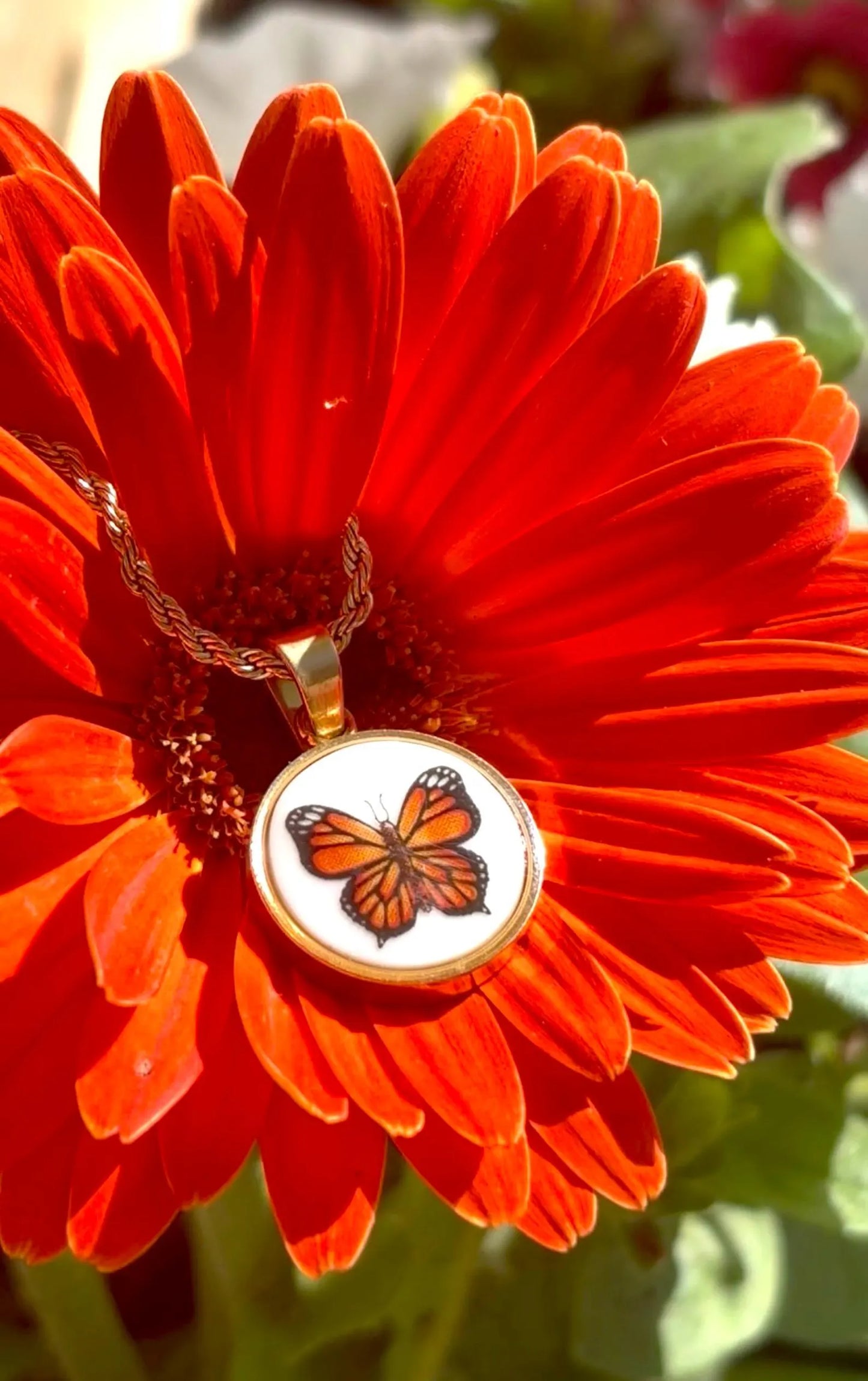 A gold-tone pendant necklace featuring an orange and black butterfly on a white background, resting on the center of a bright red gerbera daisy flower.
