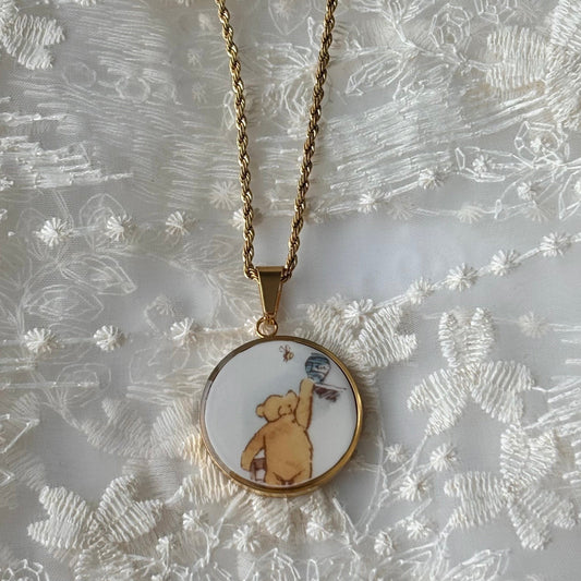 Gold necklace with a round pendant featuring a bear illustration on a textured white background