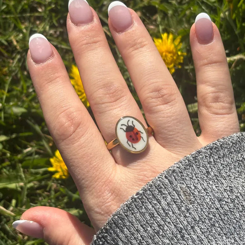 A gold signet ring featuring an oval white face with a red ladybug illustration, worn on a person's ring finger against a background of green grass and yellow dandelions.