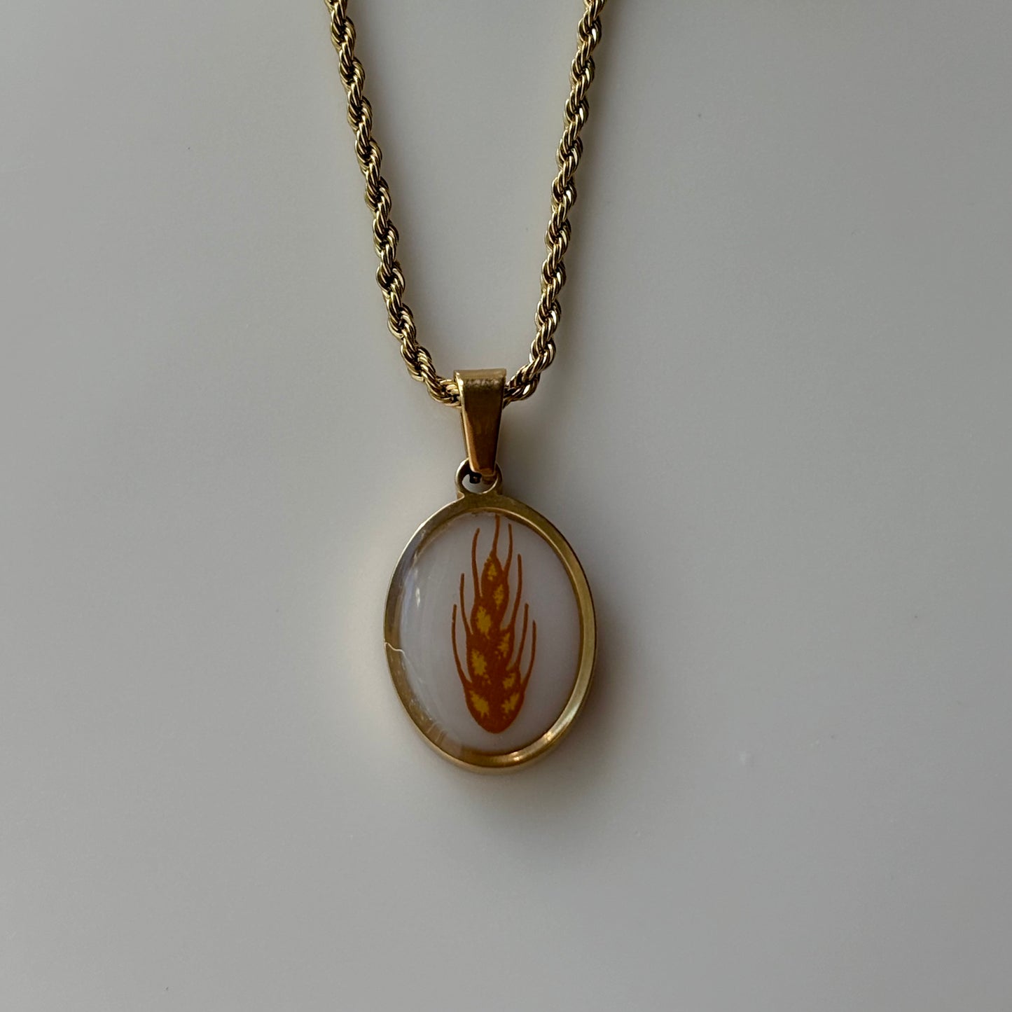 A gold chain necklace with an oval pendant featuring a white background and a gold wheat design, displayed on a neutral light gray surface.