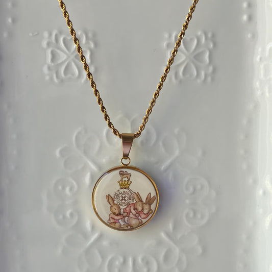 A gold-toned necklace with a round pendant featuring a vintage-style illustration of pink rabbits under a crown, displayed on a white textured background.