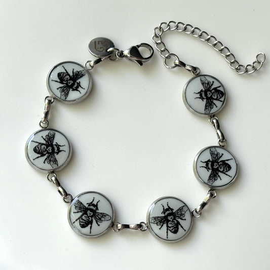 A silver-toned chain bracelet featuring five round white inlays with black printed bee designs, displayed on a clean white background.