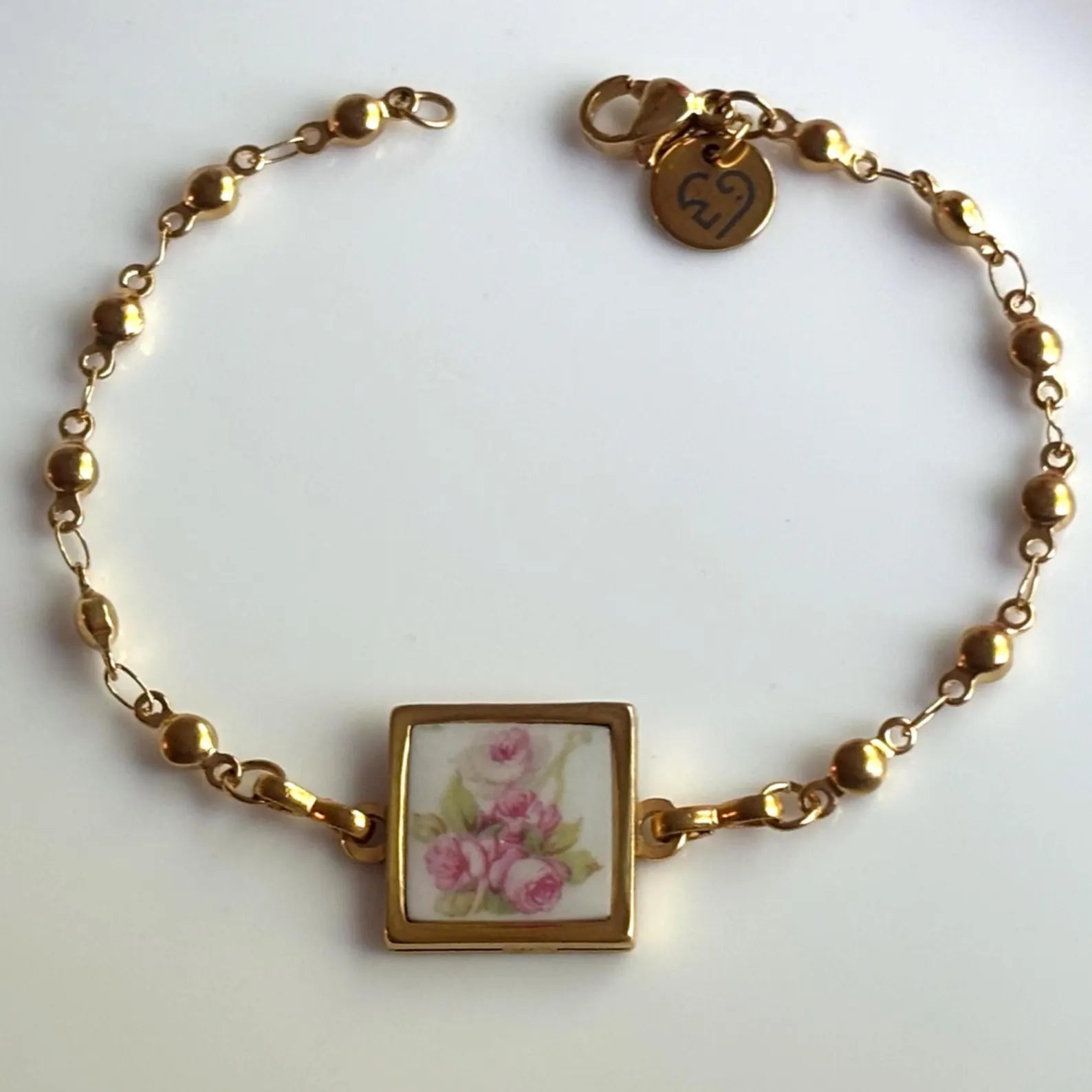 A gold chain bracelet with a square pendant featuring a pink floral design on a white background, photographed on a plain light surface.