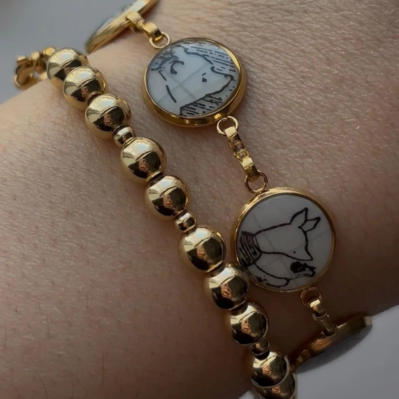 A close-up of a person's wrist wearing two gold-tone bracelets. One bracelet features a row of polished round gold beads, and the other features two oval gold charms with white enamel inserts depicting black line art illustrations.